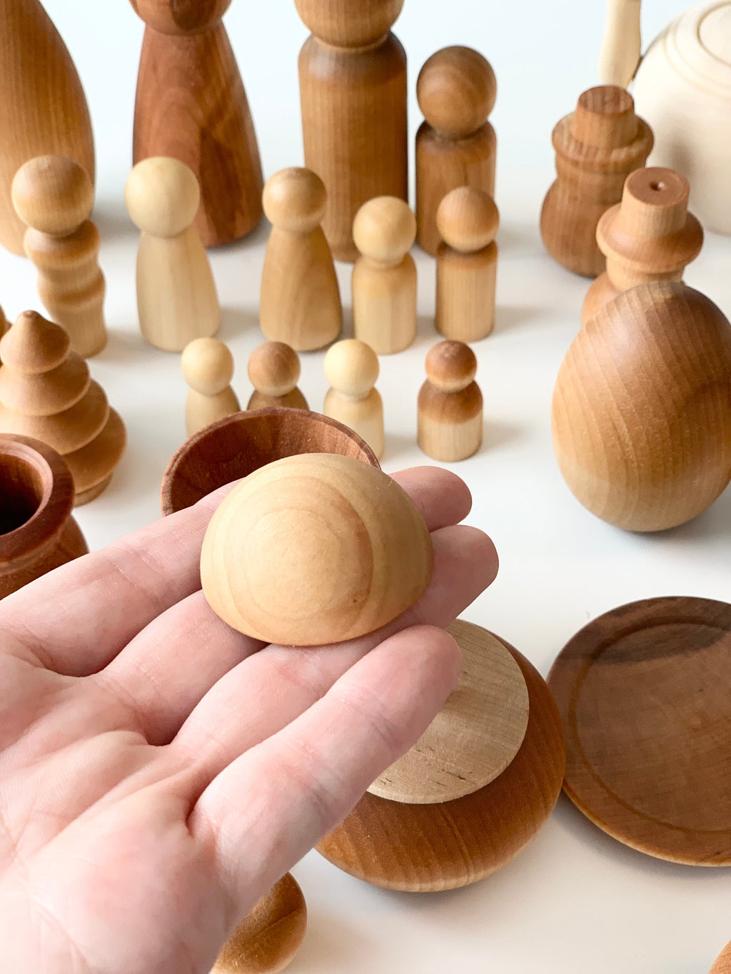 Hand holding smooth natural wood dome shape among assorted unfinished wooden figures and bowls on white surface