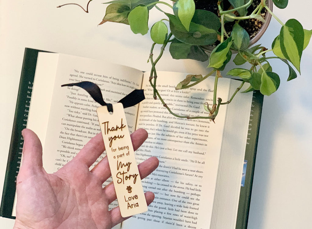 Hand holding personalized wooden bookmark with black ribbon on open book beside green potted plant on white surface
