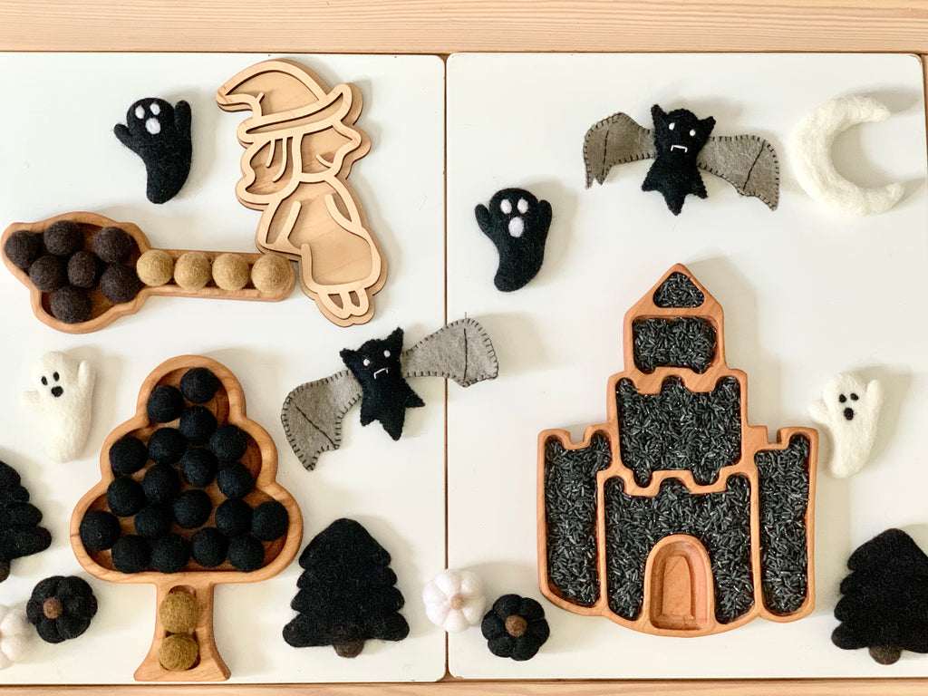 Fillable witch play tray with felt bats, ghosts, moon, tree shapes, and pom-poms on white wooden surface