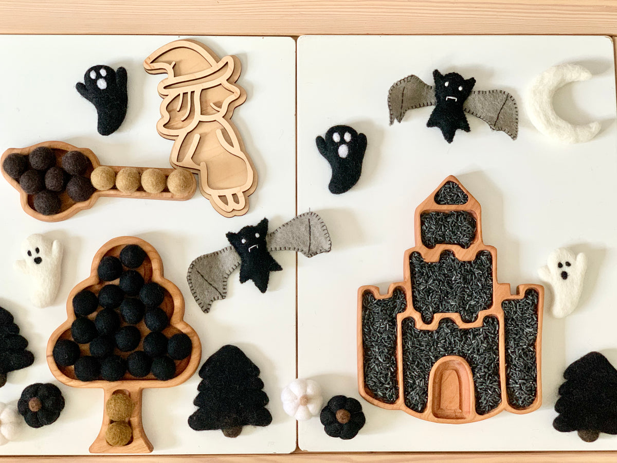 Fillable witch play tray with felt bats, ghosts, moon, tree shapes, and pom-poms on white wooden surface