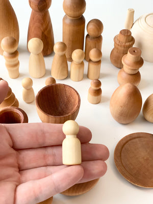 Hand holding small light natural wooden peg figure with various unfinished wooden shapes and vessels on white surface