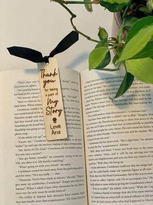 Wooden personalized bookmark with black ribbon tied at open book next to green plant on white surface