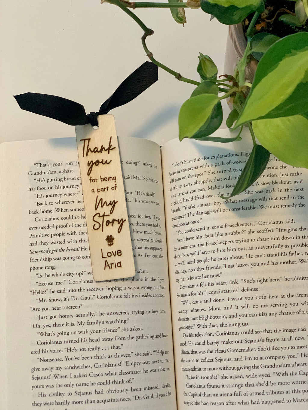 Wooden personalized bookmark with black ribbon tied at open book next to green plant on white surface