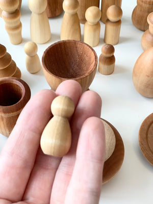 Hand holding small smooth natural wooden peg figure with various wooden bowls and peg dolls on white surface