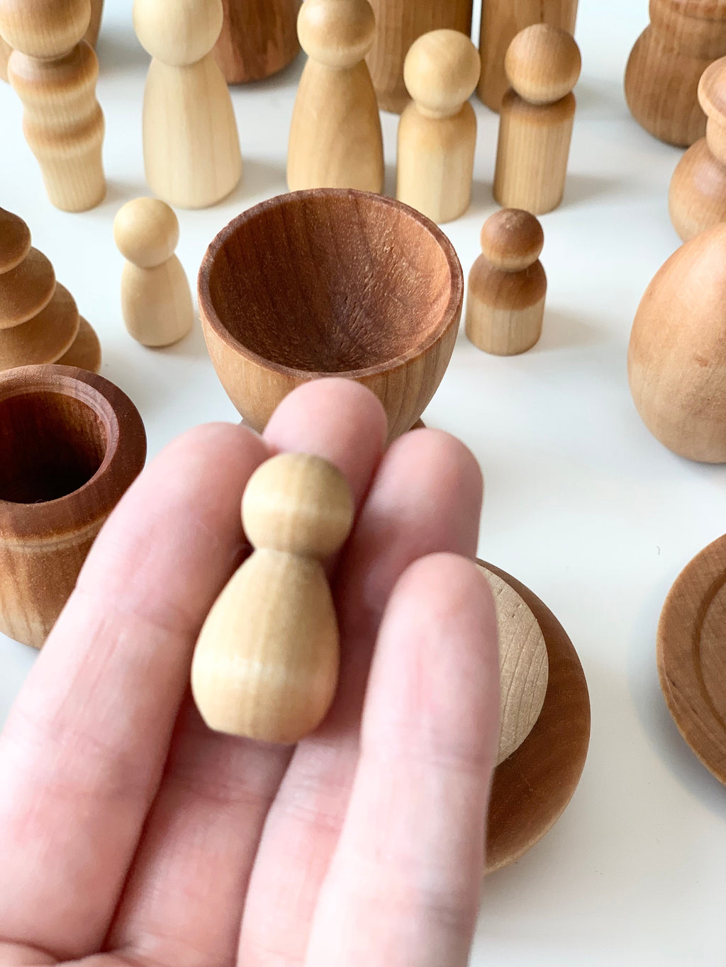 Hand holding small smooth natural wooden peg figure with various wooden bowls and peg dolls on white surface