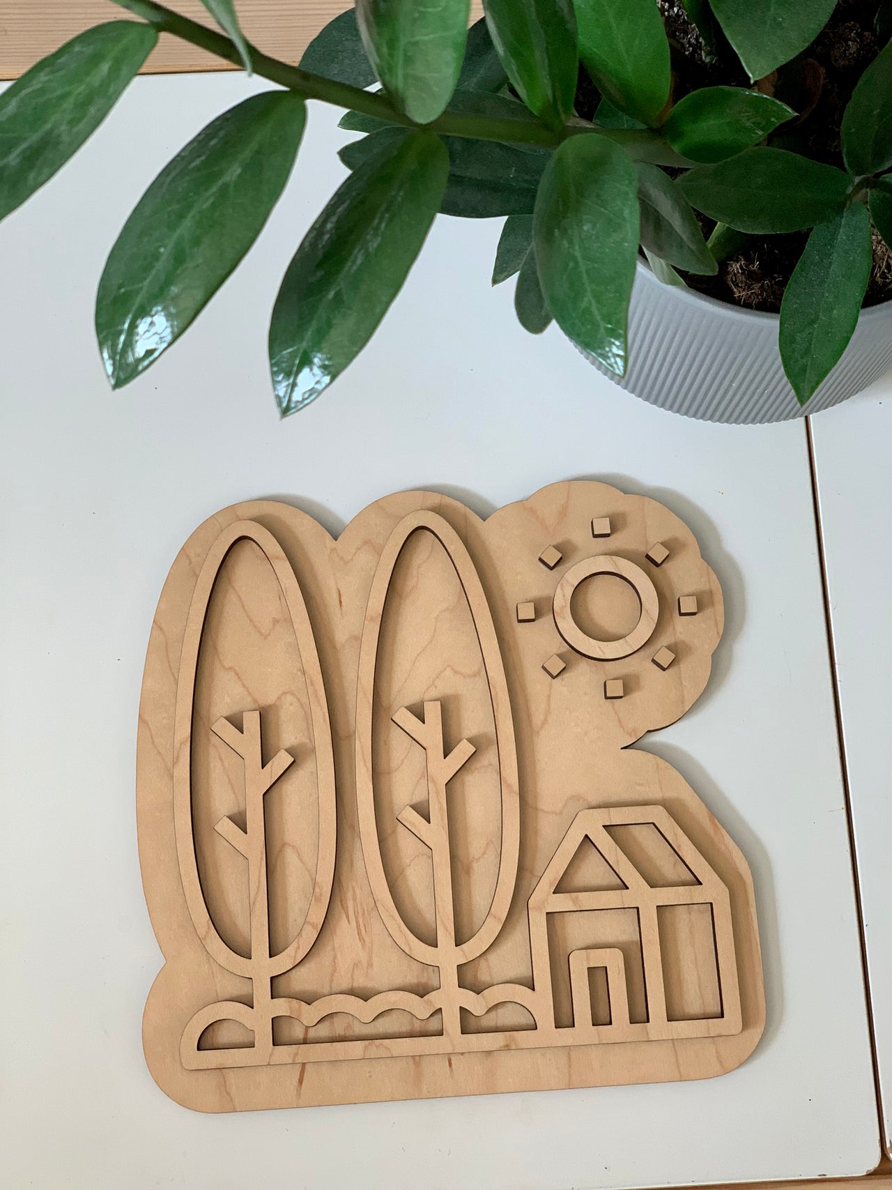 Maple wood play tray shaped like a house with trees and sun design on white table near green plant