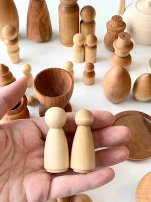 Hand holding two small natural wood peg dolls with assorted smooth wooden shapes and vessels on white surface