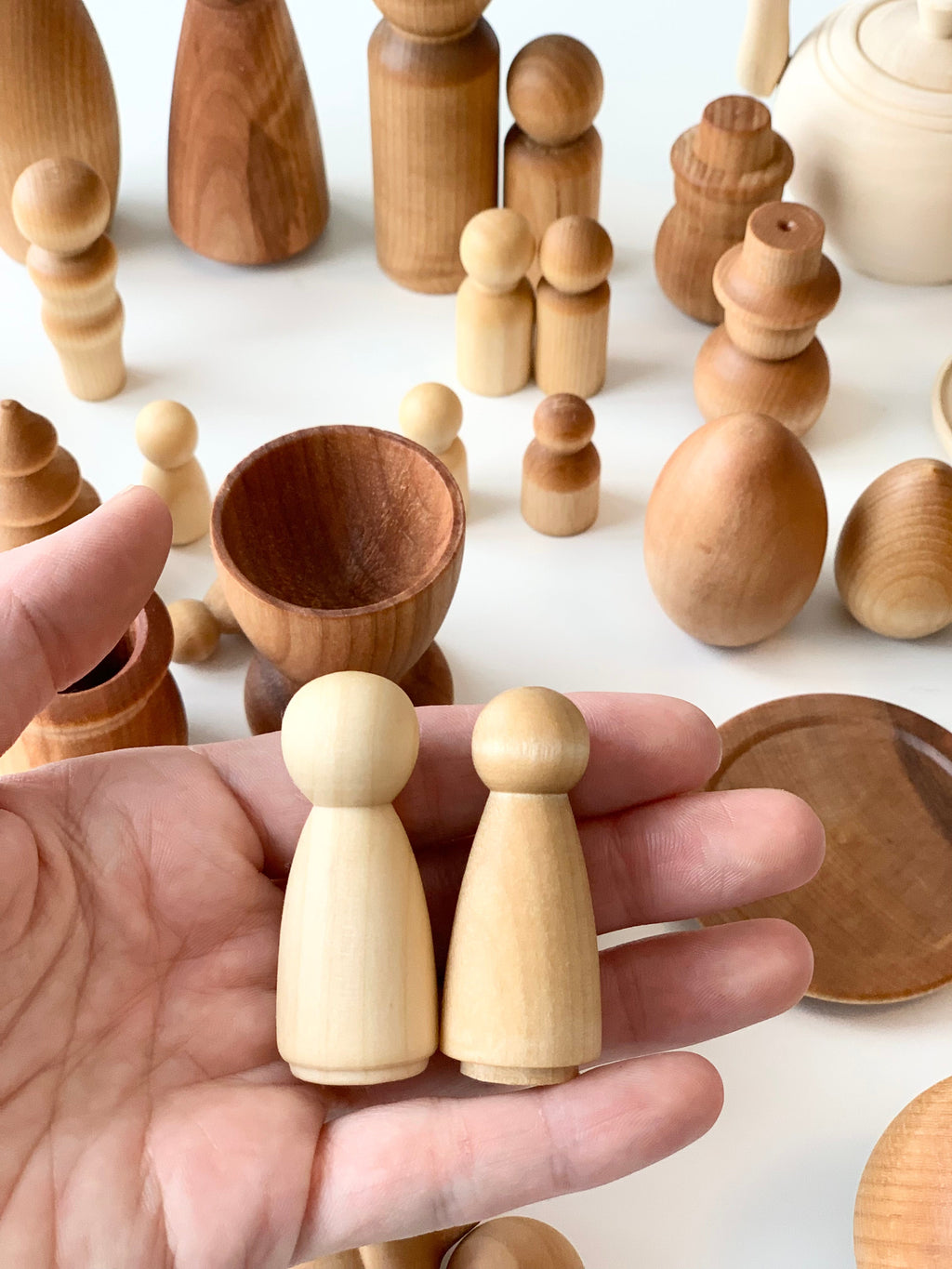 Hand holding two small natural wood peg dolls with assorted smooth wooden shapes and vessels on white surface