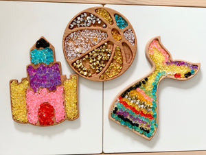 Wooden sensory trays shaped like a beach ball, castle, and mermaid tail filled with colorful translucent beads on white surface