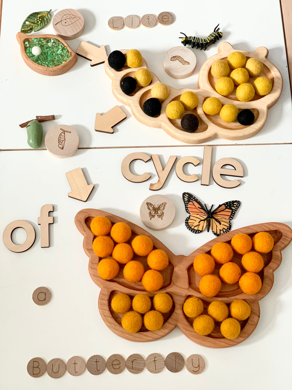 Wooden butterfly-shaped sensory tray with yellow and orange felt balls, wooden letters, arrows, and butterfly model on white surface