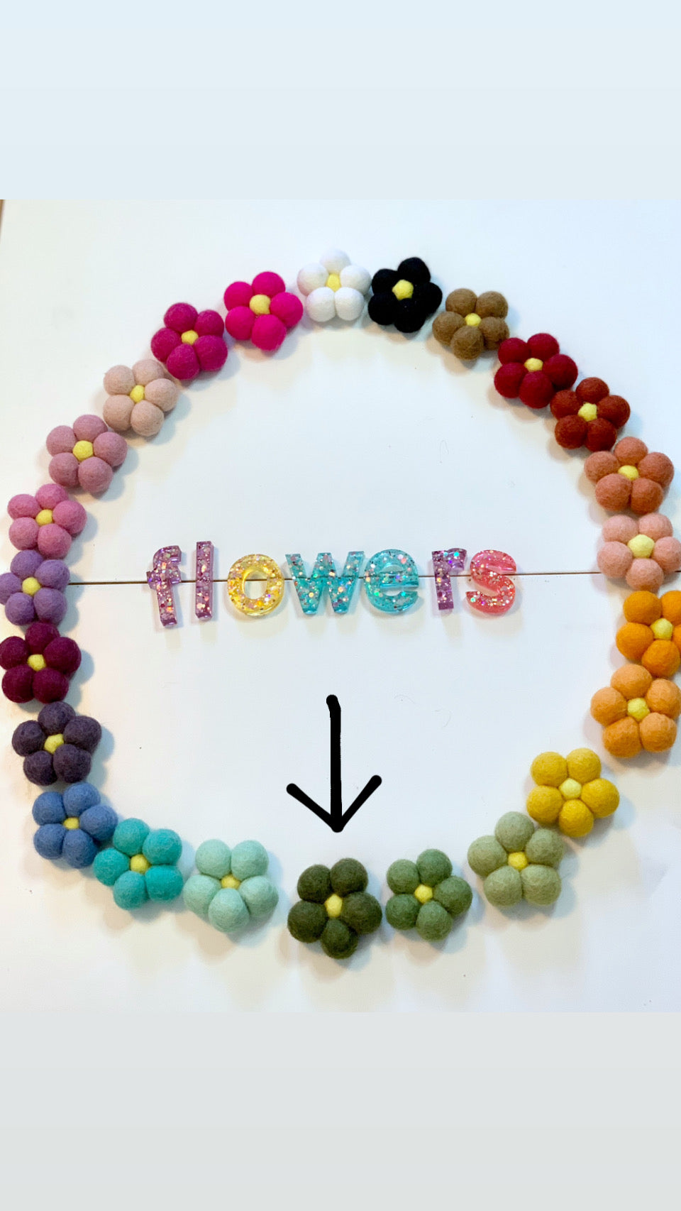 Circle of colorful handmade felt flowers with yellow centers surrounding glittery resin letters spelling flowers on white background