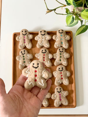 Handmade felt gingerbread person with red buttons and white icing details held over wooden tray with similar gingerbread figures on white surface