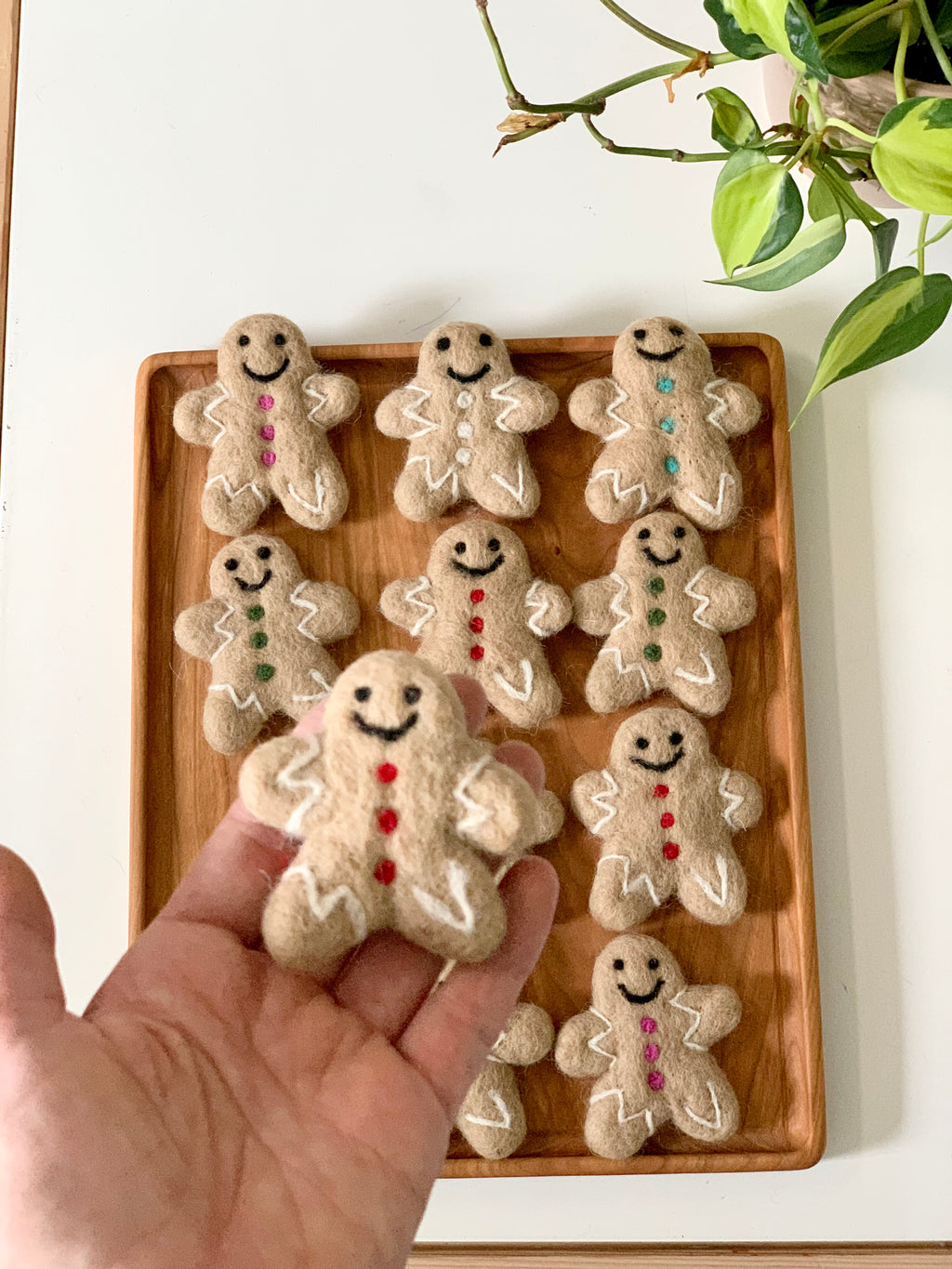 Handmade felt gingerbread person with red buttons and white icing details held over wooden tray with similar gingerbread figures on white surface