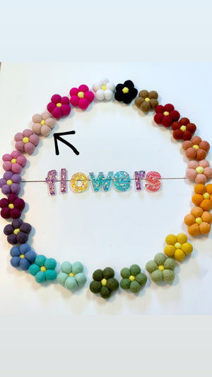 Colorful handmade felt flowers arranged in a circle surrounding glittery letters spelling flowers on white surface