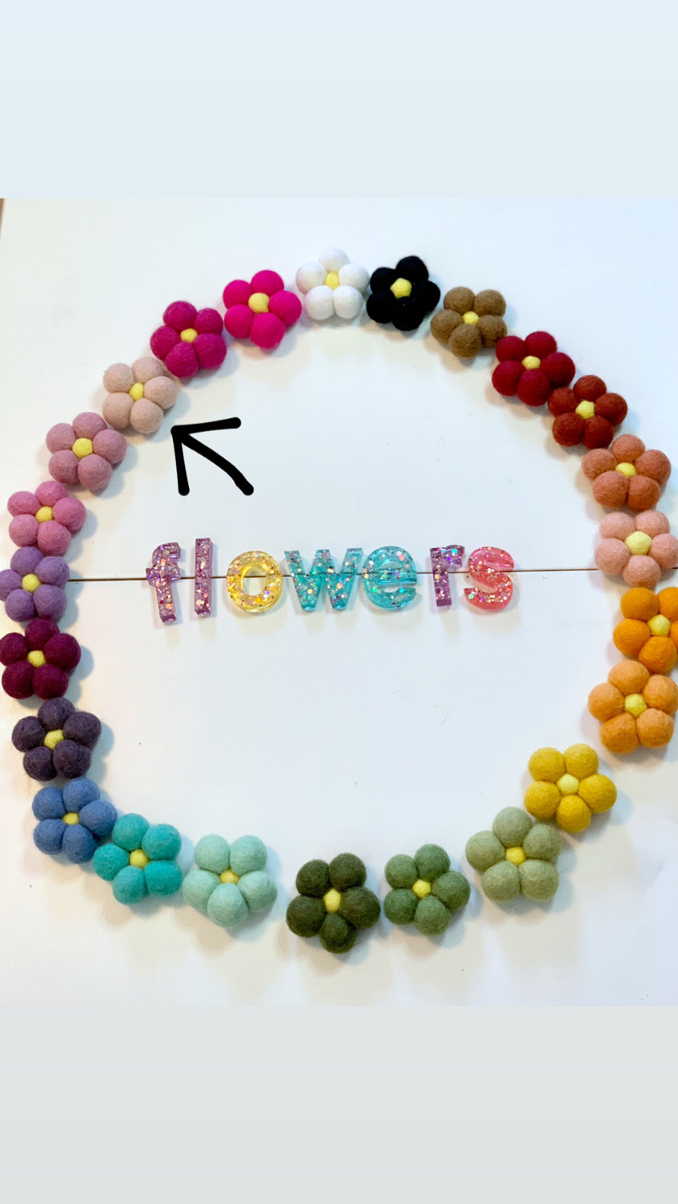 Colorful handmade felt flowers arranged in a circle surrounding glittery letters spelling flowers on white surface
