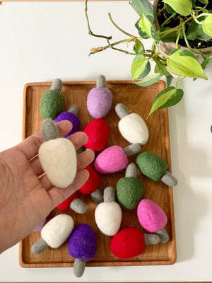 Hand holding one white felt lightbulb above wooden tray with scattered felt lightbulbs in multiple colors and plant nearby