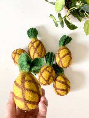 Handmade felt pineapple with green leaves held by hand surrounded by five similar felt pineapples on white surface