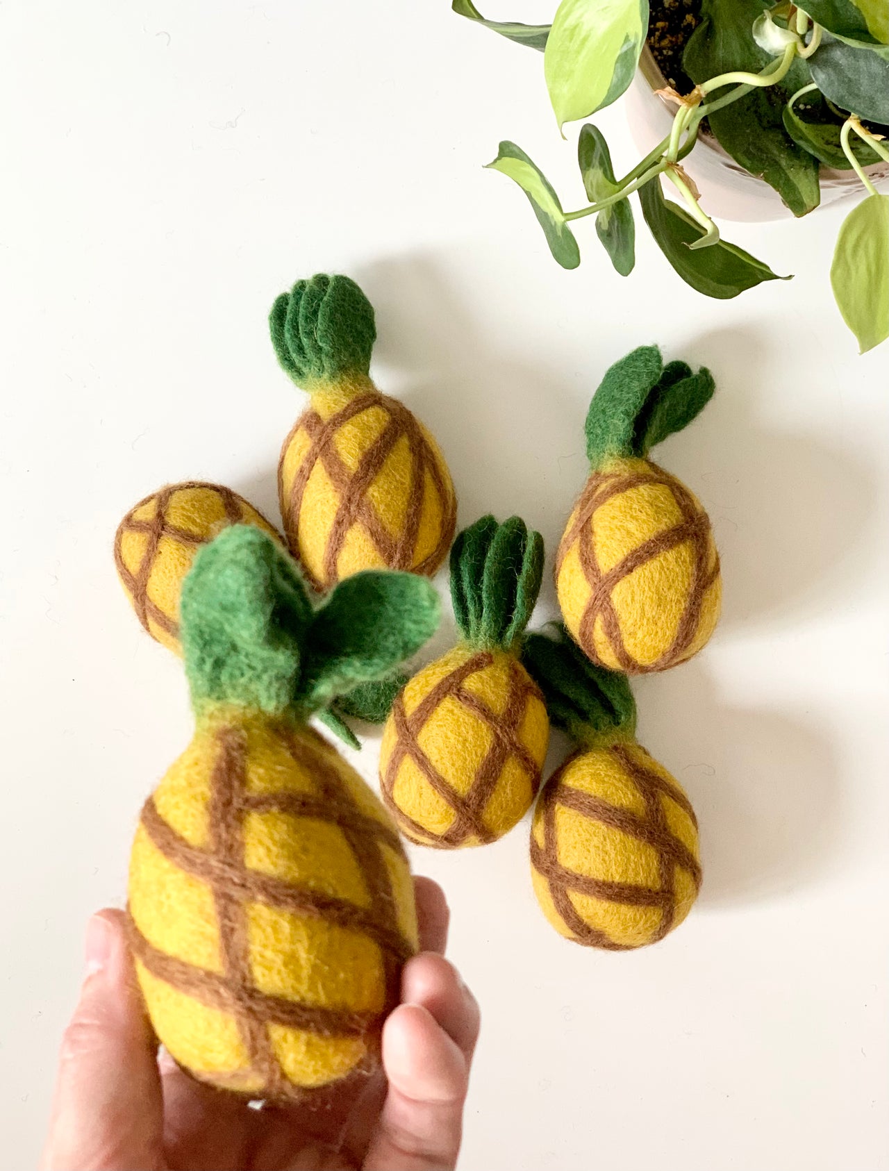 Handmade felt pineapple with green leaves held by hand surrounded by five similar felt pineapples on white surface