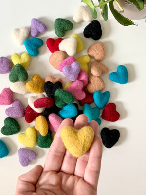 Small handmade felt heart in mustard yellow held in hand with many colorful felt hearts scattered on white surface