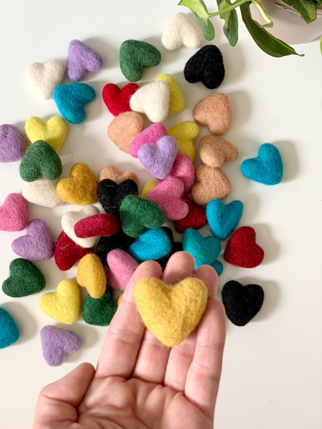 Small handmade felt heart in mustard yellow held in hand with many colorful felt hearts scattered on white surface