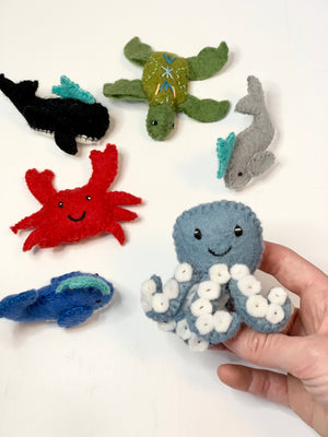 Handmade felt blue octopus with white suction cups held by hand surrounded by other felt sea creatures on white background