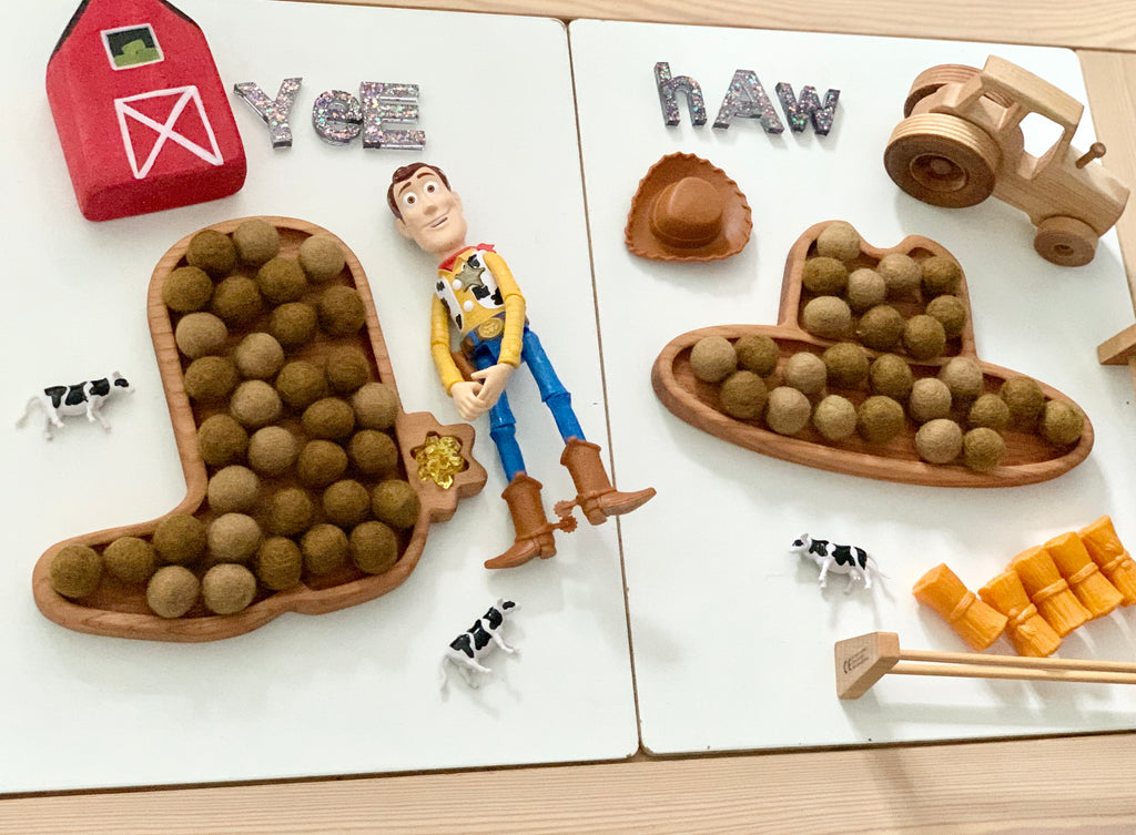 Wooden cowboy boot and hat sensory trays filled with felt balls, toy tractor, cows, barn, Woody figure, and hay bales on white boards