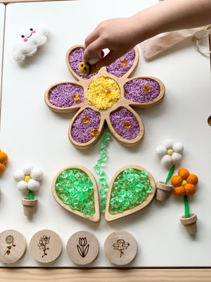 Child’s hand placing a bee toy on a wooden flower tray filled with colored rice and surrounded by felt flowers and game pieces