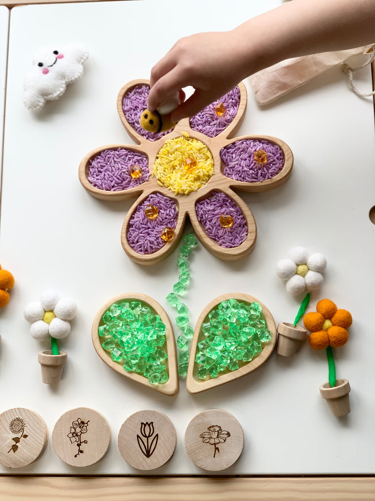 Child’s hand placing a bee toy on a wooden flower tray filled with colored rice and surrounded by felt flowers and game pieces