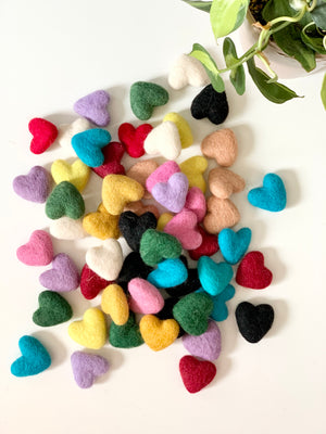 Assorted small handmade felt hearts in various colors scattered on white surface with green plant nearby