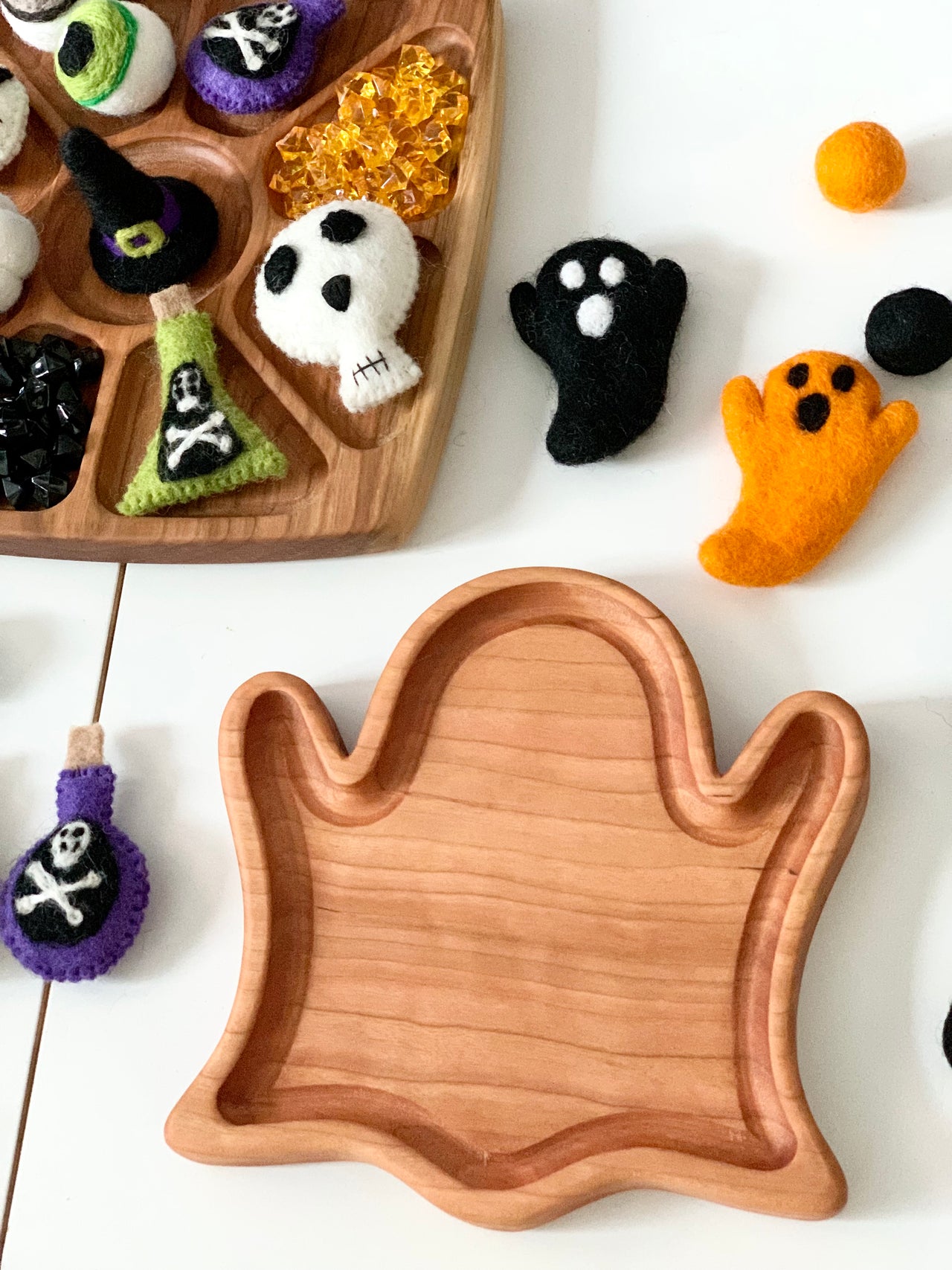 Wooden ghost-shaped sensory tray surrounded by colorful felt ghosts, skull, potions, and amber gems on white surface
