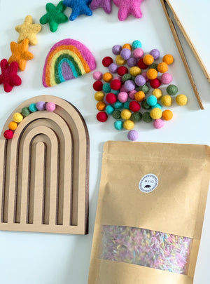 Laser cut wooden rainbow tray with colorful felt balls, felt rainbow, star decorations, wooden tweezers, and craft supplies on white table