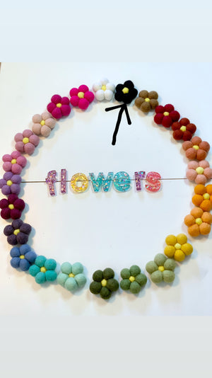 Single black felt flower with yellow center among colorful felt flowers arranged in a circle on white background