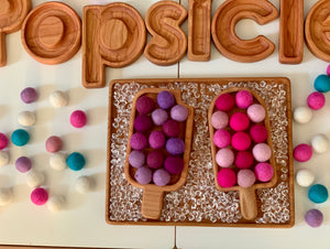 Wooden popsicle-shaped sensory trays filled with colorful felt balls on textured wooden board with wooden letter tiles spelling popsicle