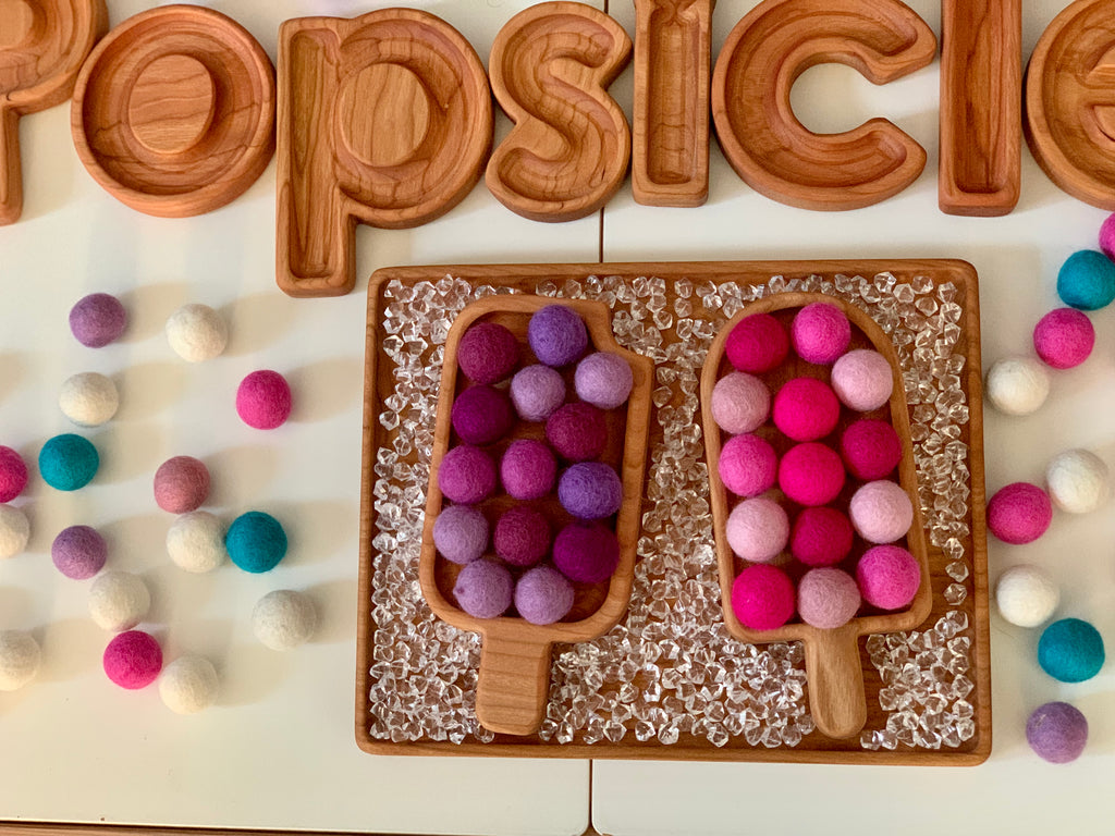Wooden popsicle-shaped sensory trays filled with colorful felt balls on textured wooden board with wooden letter tiles spelling popsicle