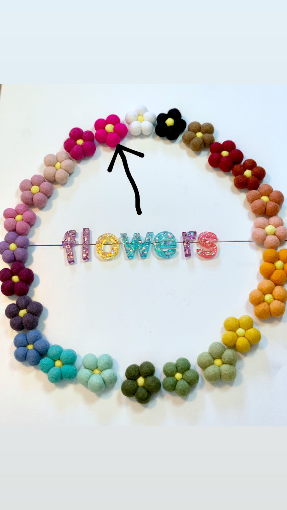 Single white handmade felt flower with yellow center among colorful felt flowers arranged in circle on white background