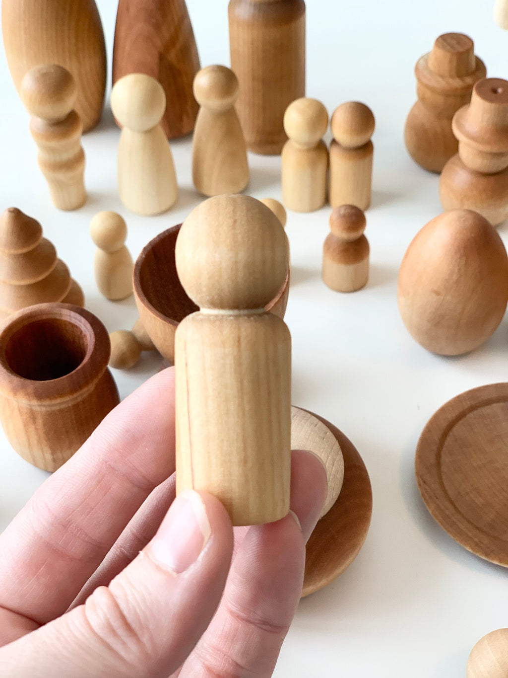 Hand holding smooth light wood peg figure with various natural wood shapes and vessels in soft lighting on white surface