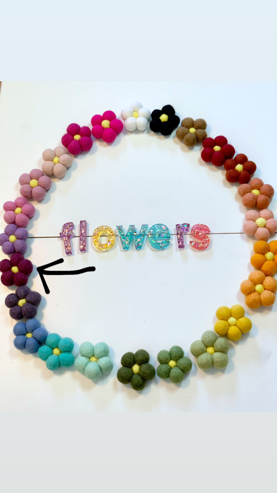 Colorful handmade felt flowers arranged in a circle around glittery resin letters spelling flowers on white background