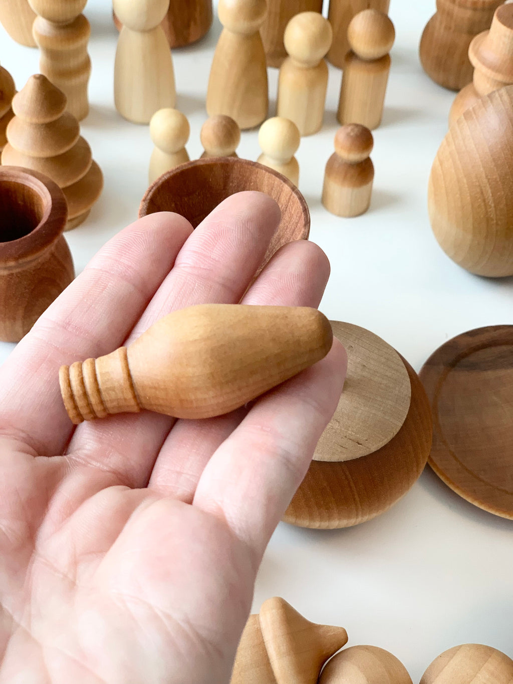 Hand holding smooth natural wooden utensil shape with various wooden figures and bowls on white surface in background
