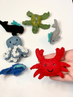 Handmade red felt crab with smiling face held in hand alongside other colorful sea creature felt toys on white surface