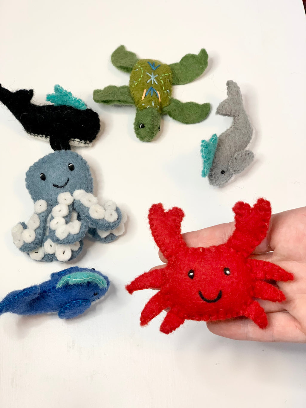 Handmade red felt crab with smiling face held in hand alongside other colorful sea creature felt toys on white surface