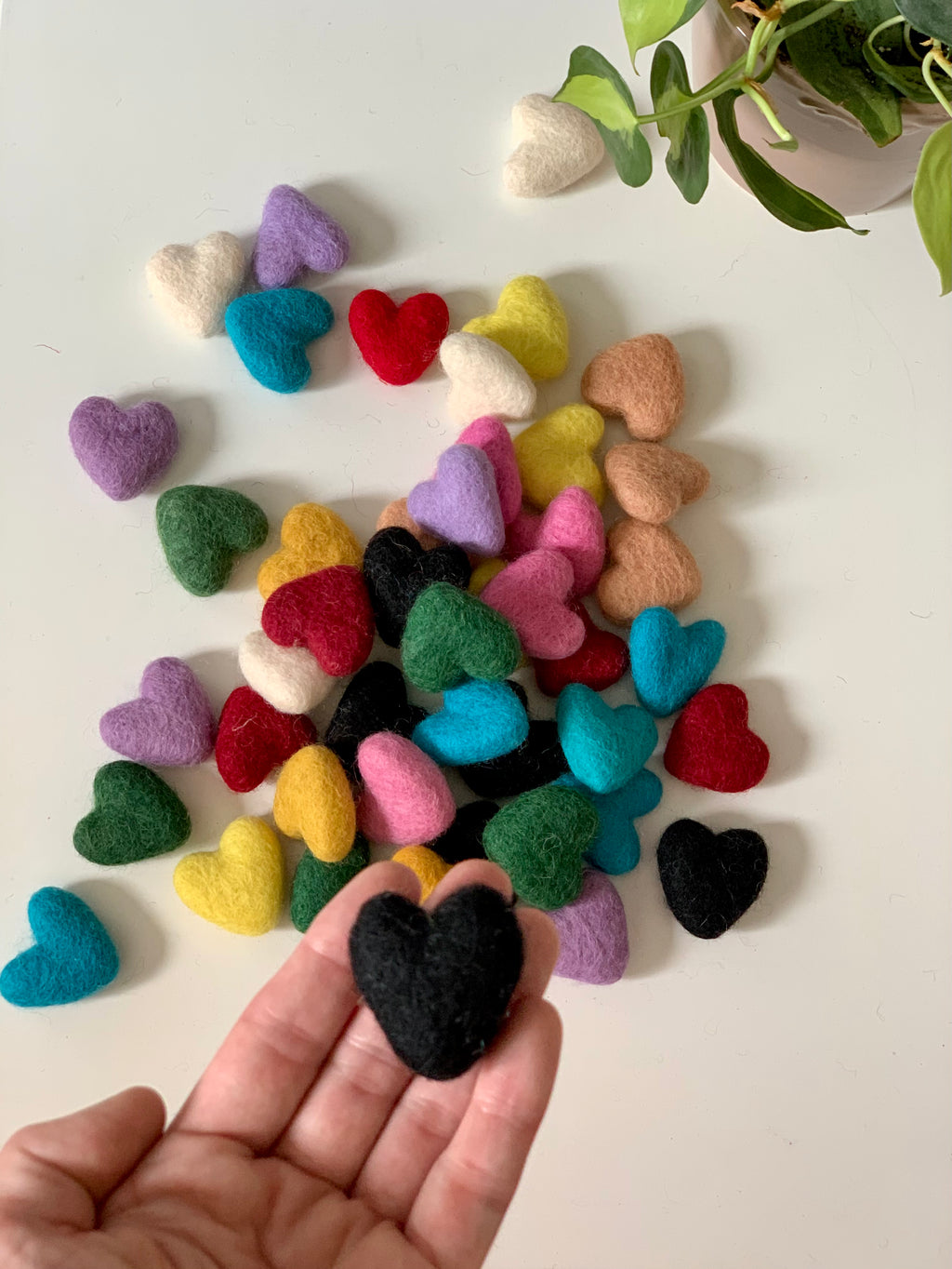 Assorted handmade small felt hearts in various colors with one black heart held in foreground on white surface