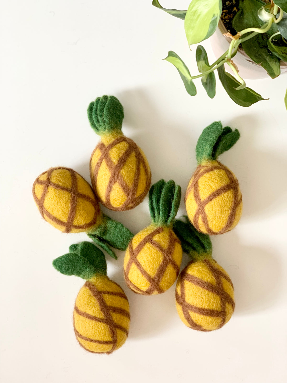 Group of six handmade felt pineapples with green tops on white surface next to potted plant