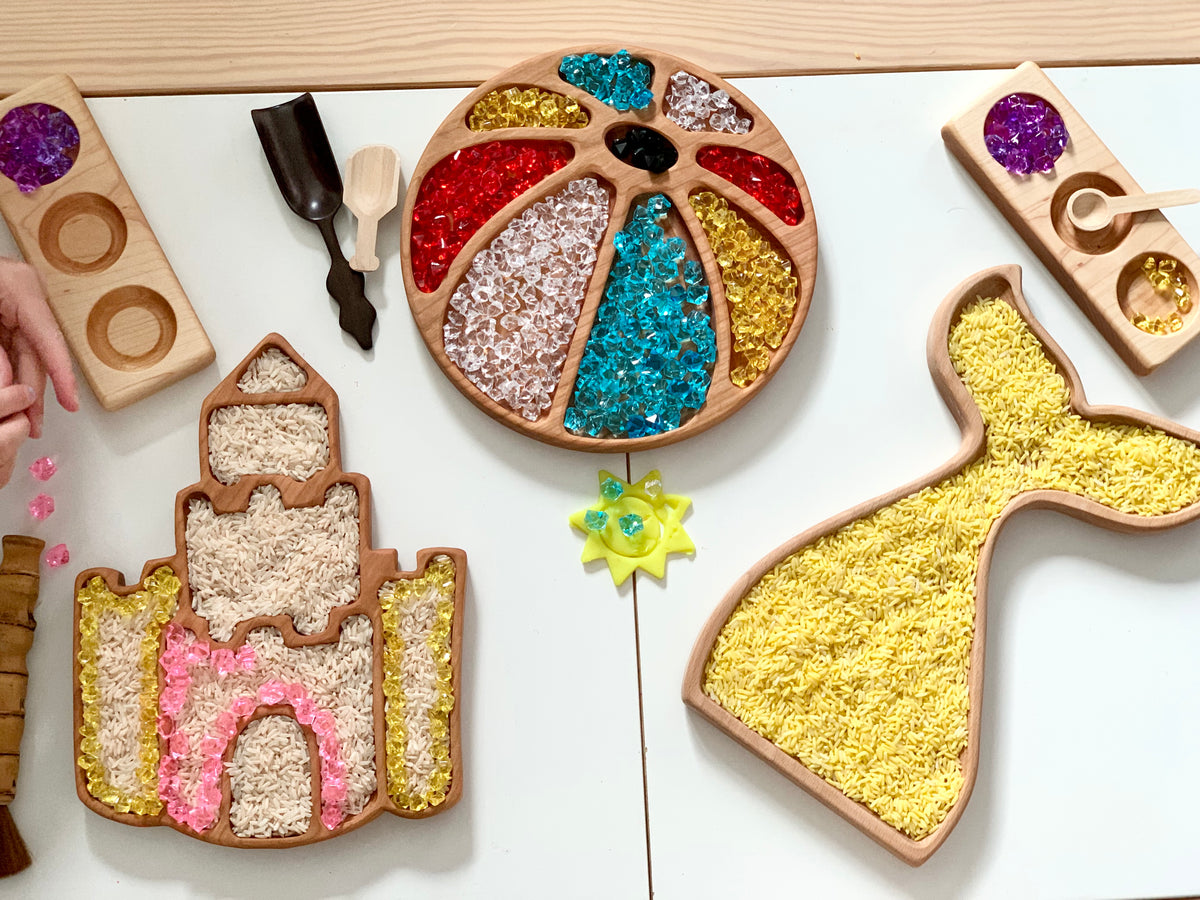 Wooden sensory trays shaped like a sandcastle, beach ball, and whale filled with colorful sensory beads and tools on white table