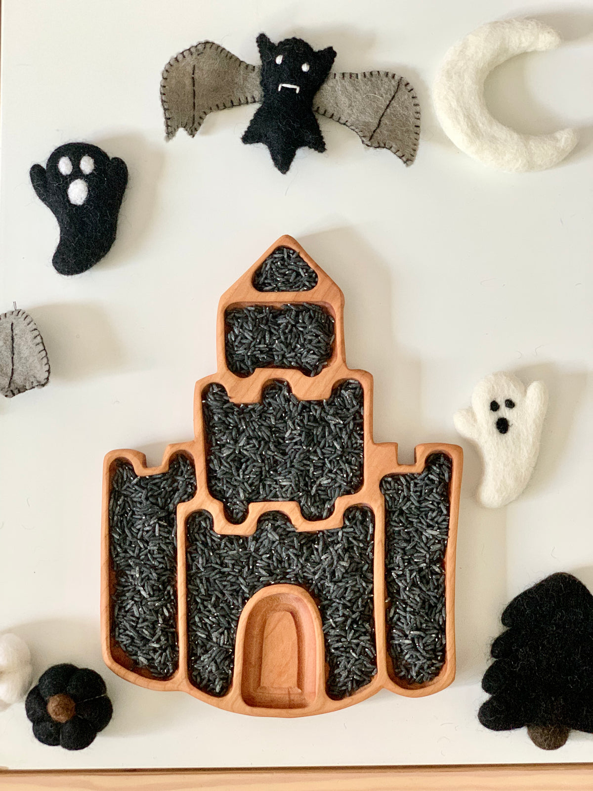 Castle-shaped wooden sensory tray filled with black rice surrounded by felt bats, ghosts, crescent moon, and pumpkin on white surface