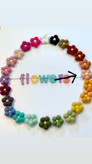 Circle of small handmade felt flowers in various colors surrounding colorful glittery letters spelling flowers on white background