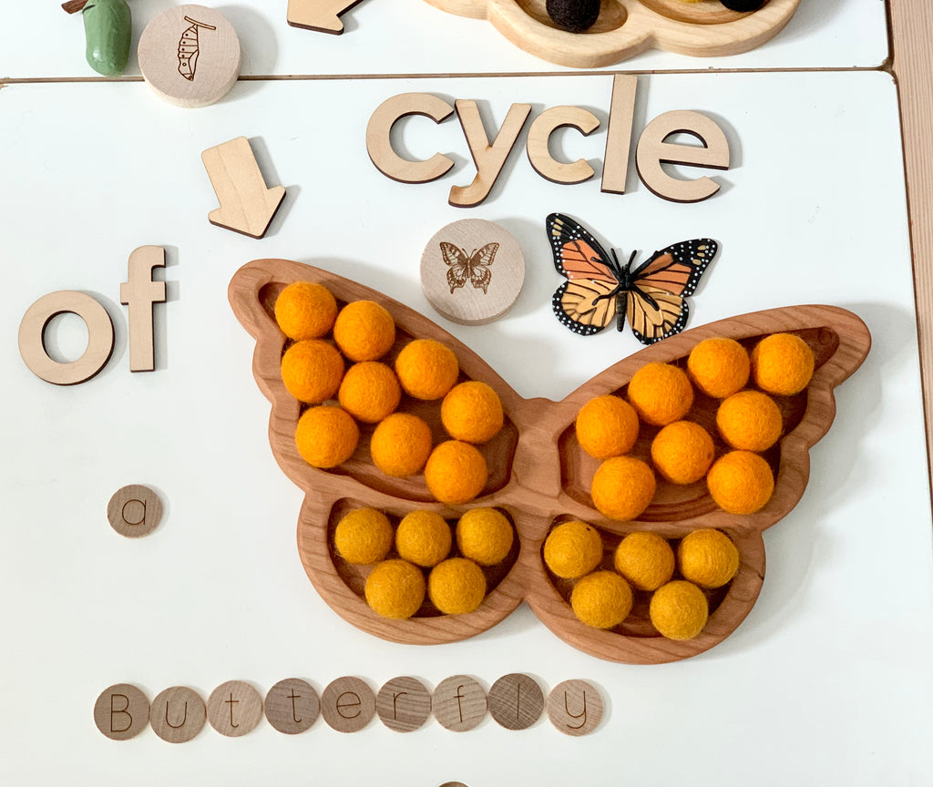 Wooden butterfly-shaped sensory tray with orange and yellow felt balls surrounded by wooden letters and butterfly illustrations