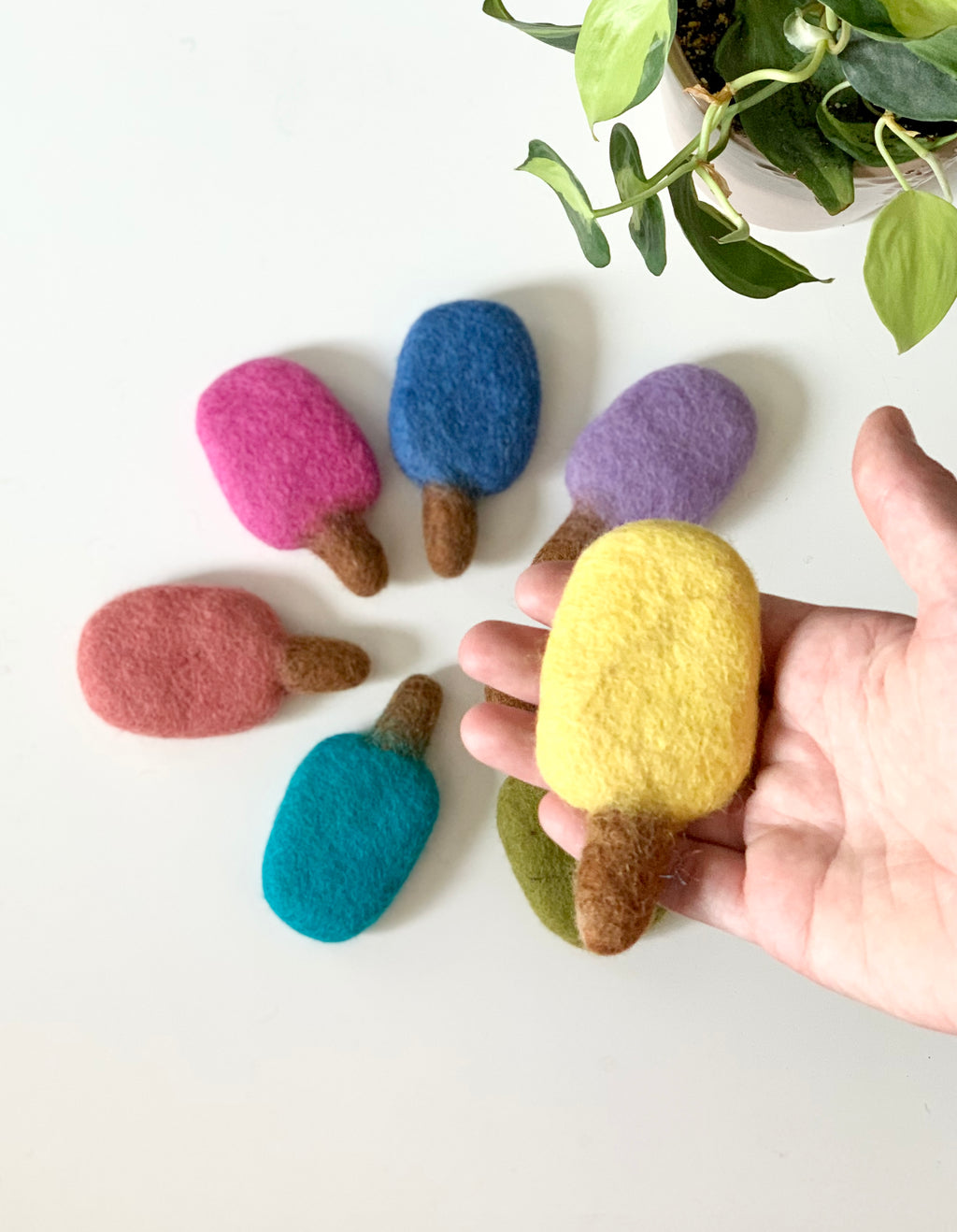 Hand holding one yellow felt popsicle with six other colorful felt popsicles on white table beside green plant
