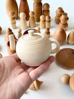 Hand holding small unfinished natural wood teapot toy with assorted smooth wooden peg dolls and bowls on white surface