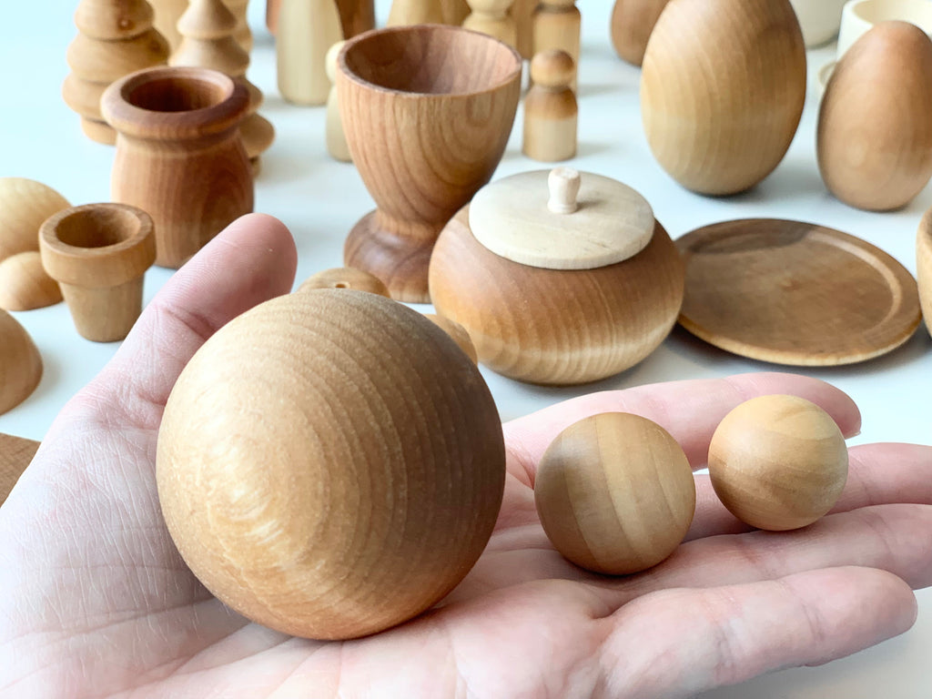 Hand holding three smooth unfinished wooden balls of varying sizes with assorted wooden shapes in the background