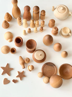 Unfinished light and dark wooden figurines, bowls, cups, stars, hearts, and a teapot arranged on a white surface